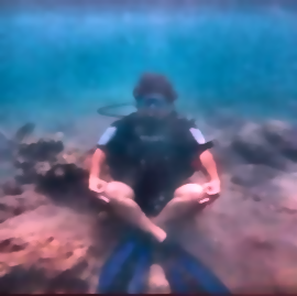 Color-restored underwater frame showing the same seated scuba diver.